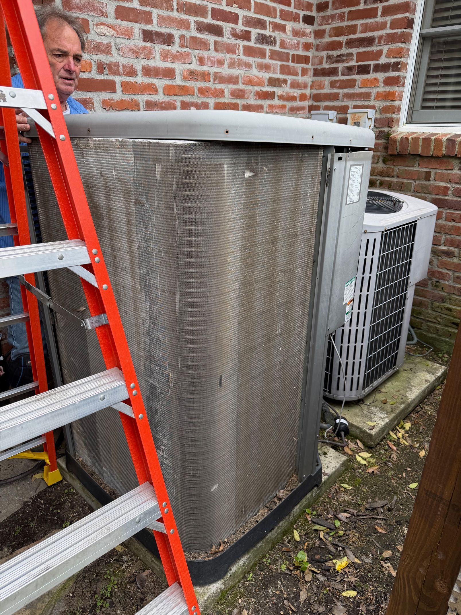 Outdoor HVAC unit beside brick wall, with a red ladder leaning against it and a person partly visible on the left.