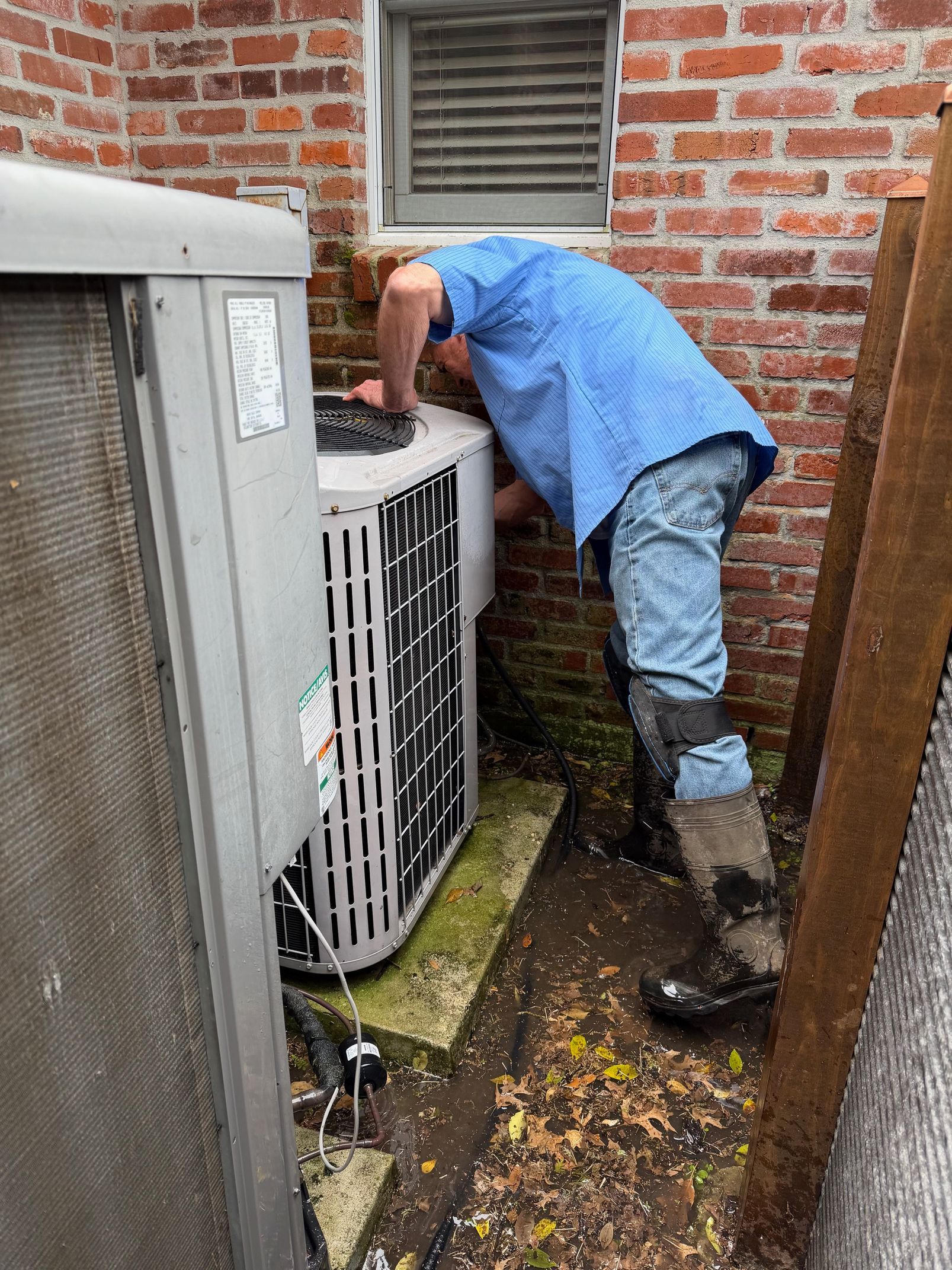 Technician inspecting an outdoor HVAC unit beside a brick wall, partly hidden between buildings.