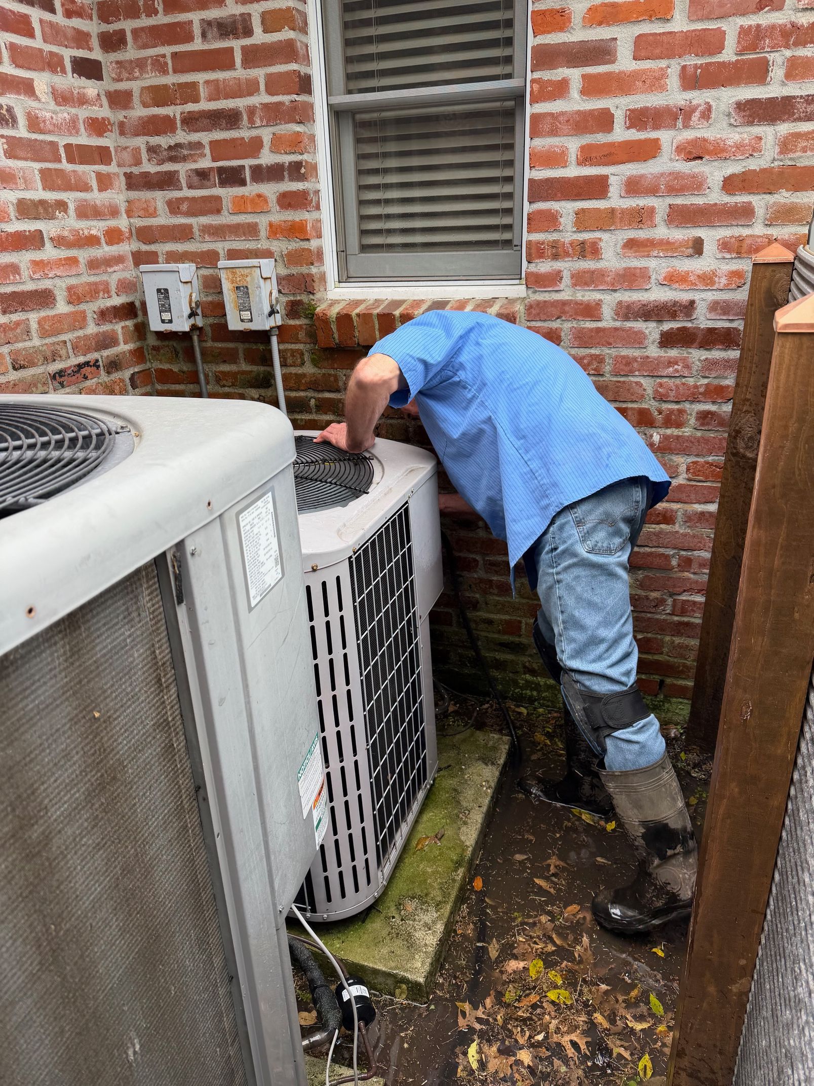 Technician in blue shirt inspecting outdoor air conditioner units against a brick wall.