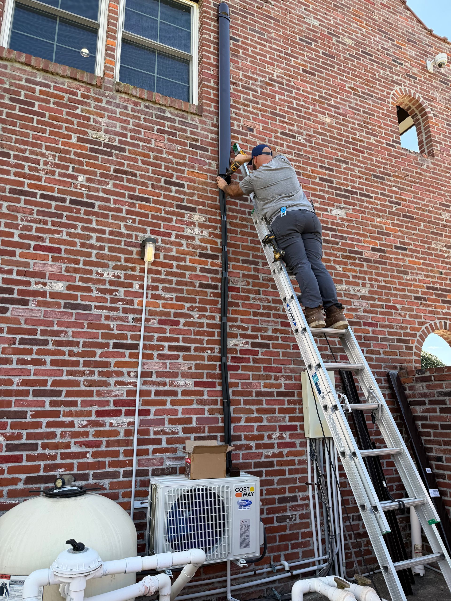 Worker on a ladder repairing a vertical pipe on a red brick building.
