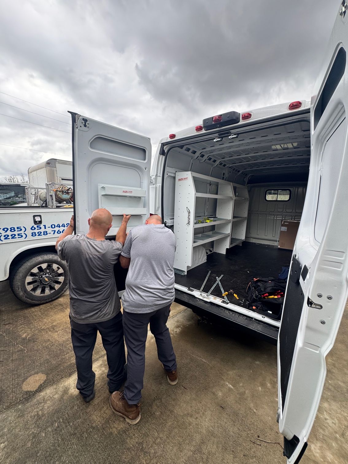 Two workers inspect the open rear of a white van in a cloudy parking lot.