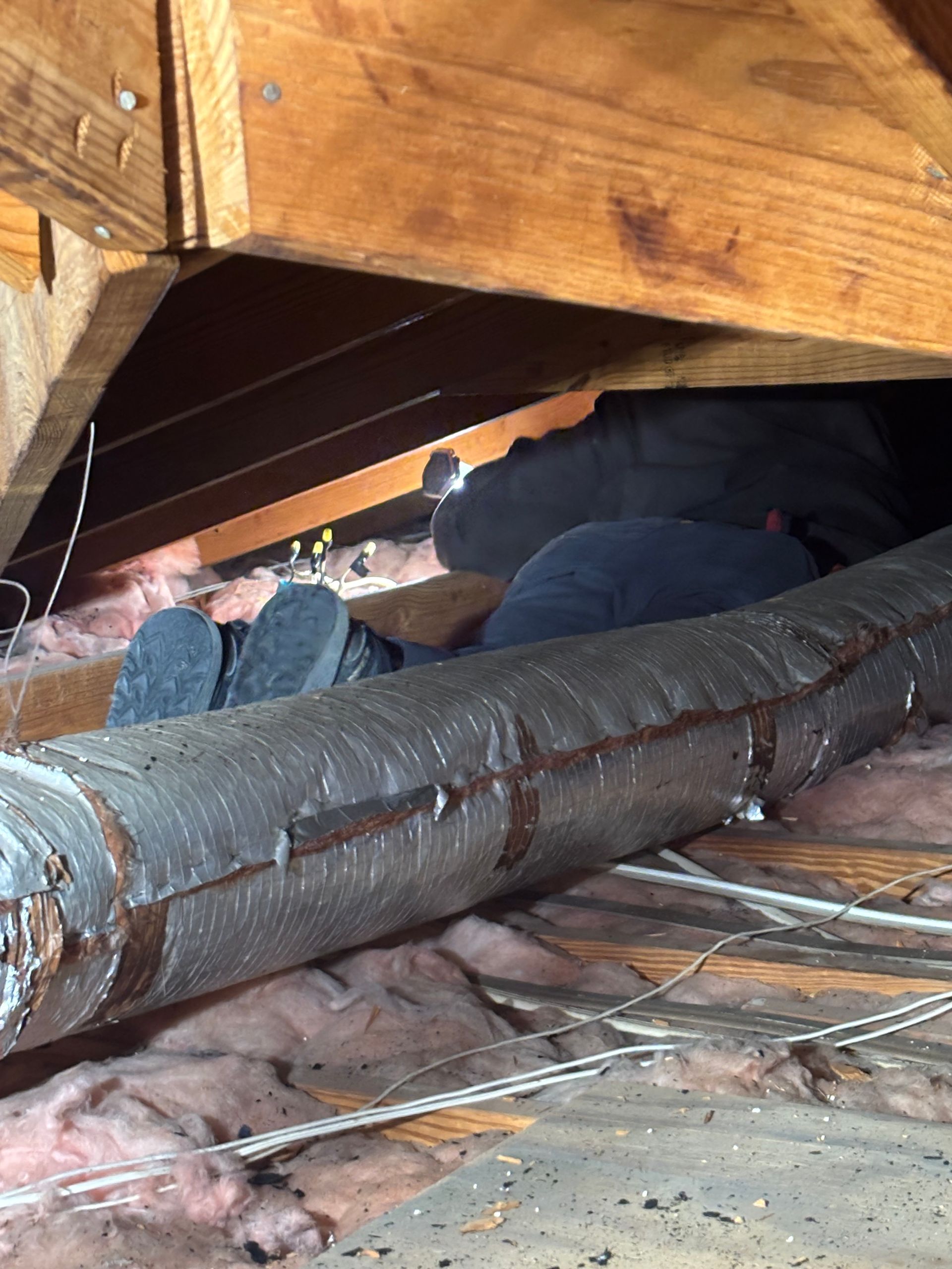 Person working in a cramped attic crawlspace near exposed wooden beams and HVAC ducting