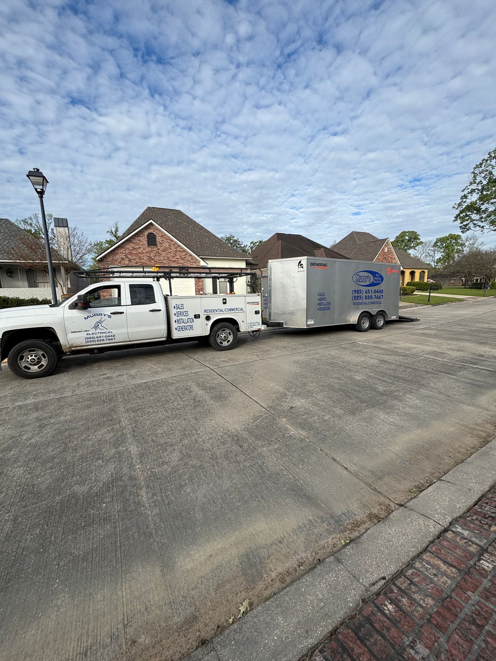 White utility truck towing a silver trailer on a gravel lot beside brick houses under a cloudy sky