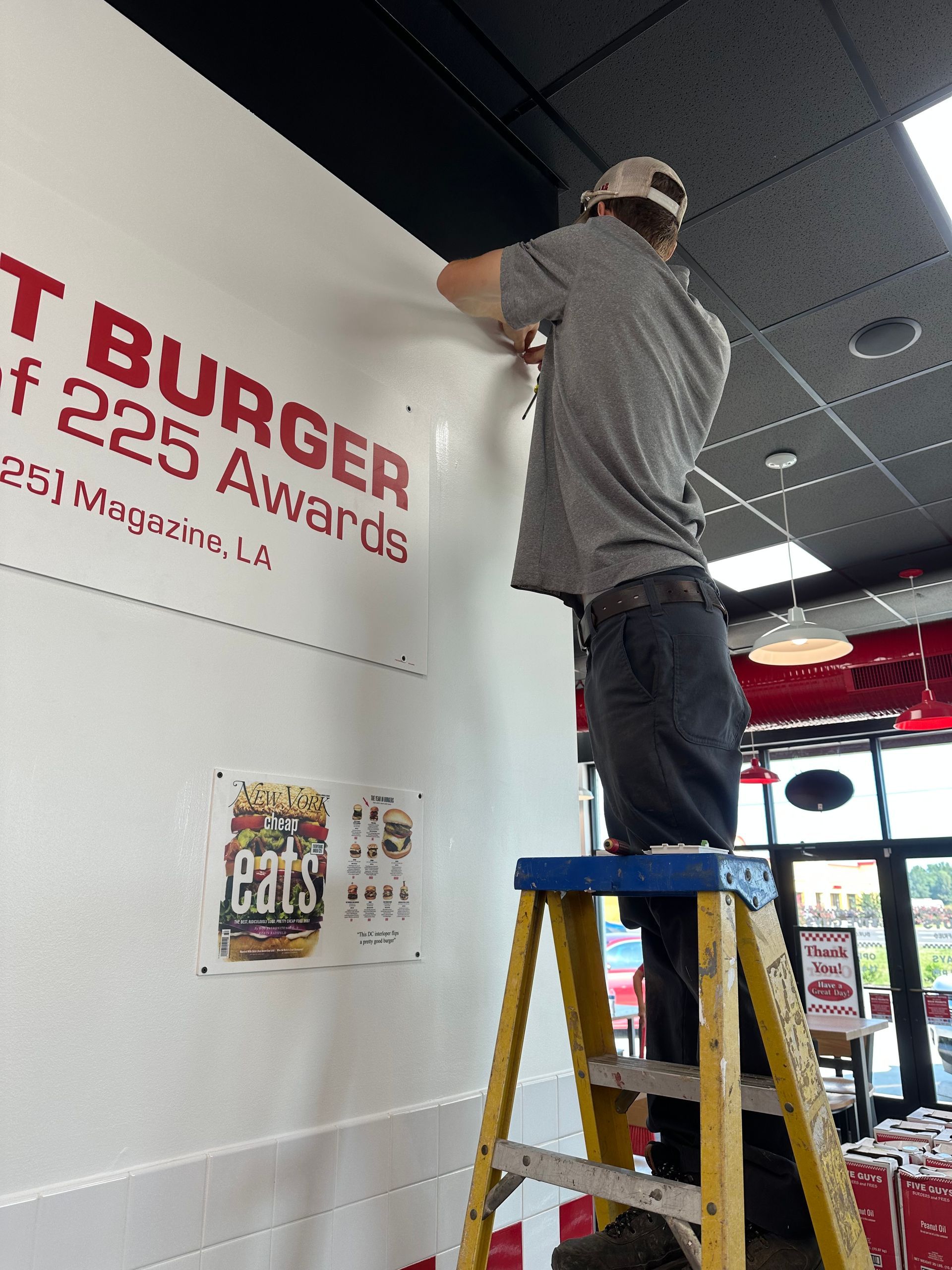 Man on ladder installing red lettering on a white wall inside a restaurant, with ceiling lights above.
