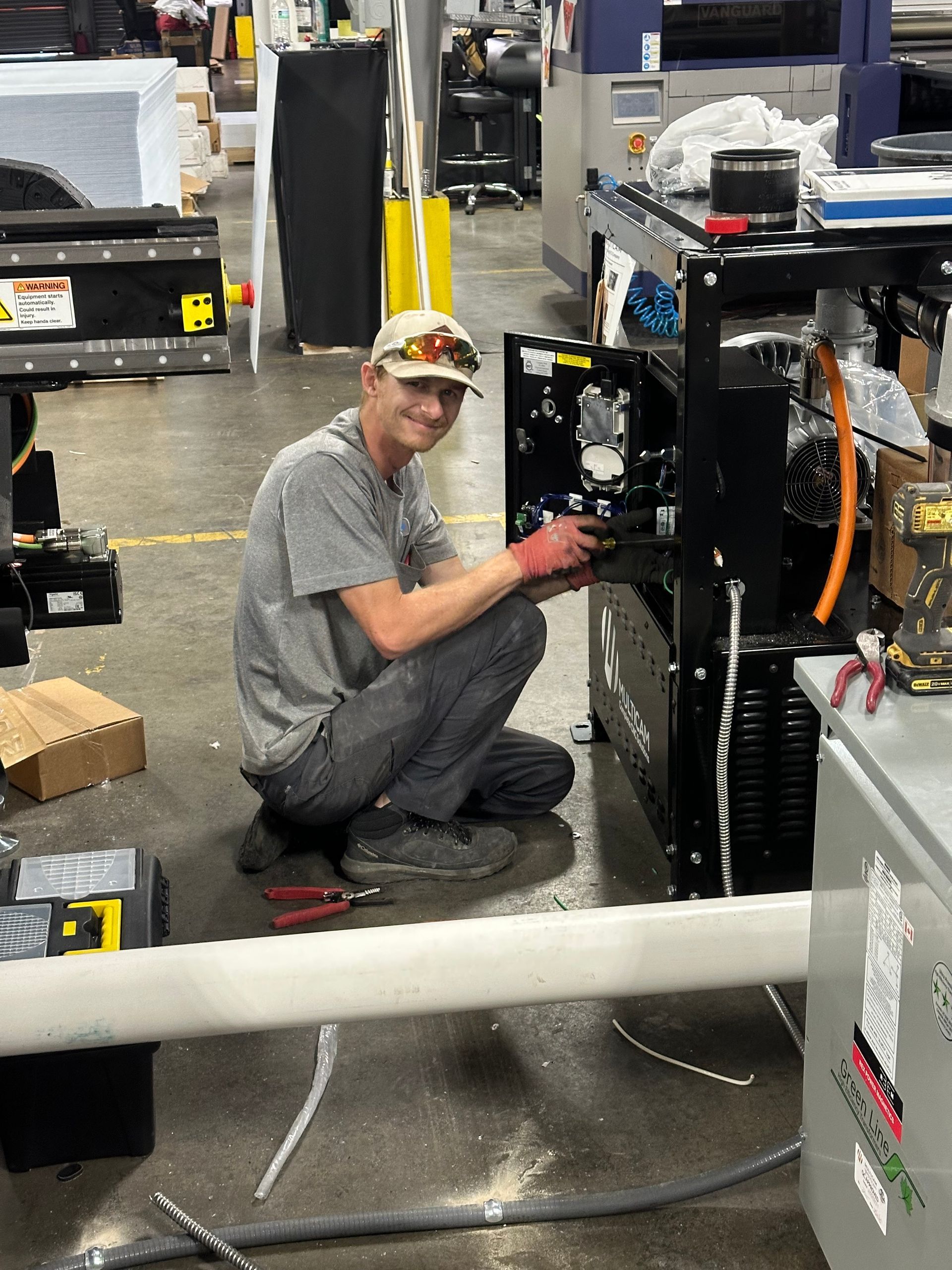Worker kneeling beside a machine in a factory, using a tool and smiling at the camera.