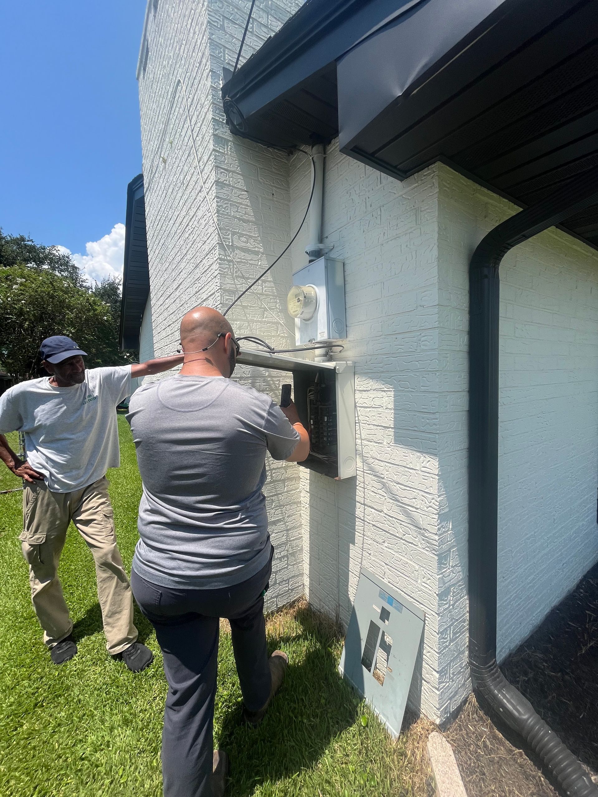 Two people inspect an exterior wall-mounted utility box on a stucco house.