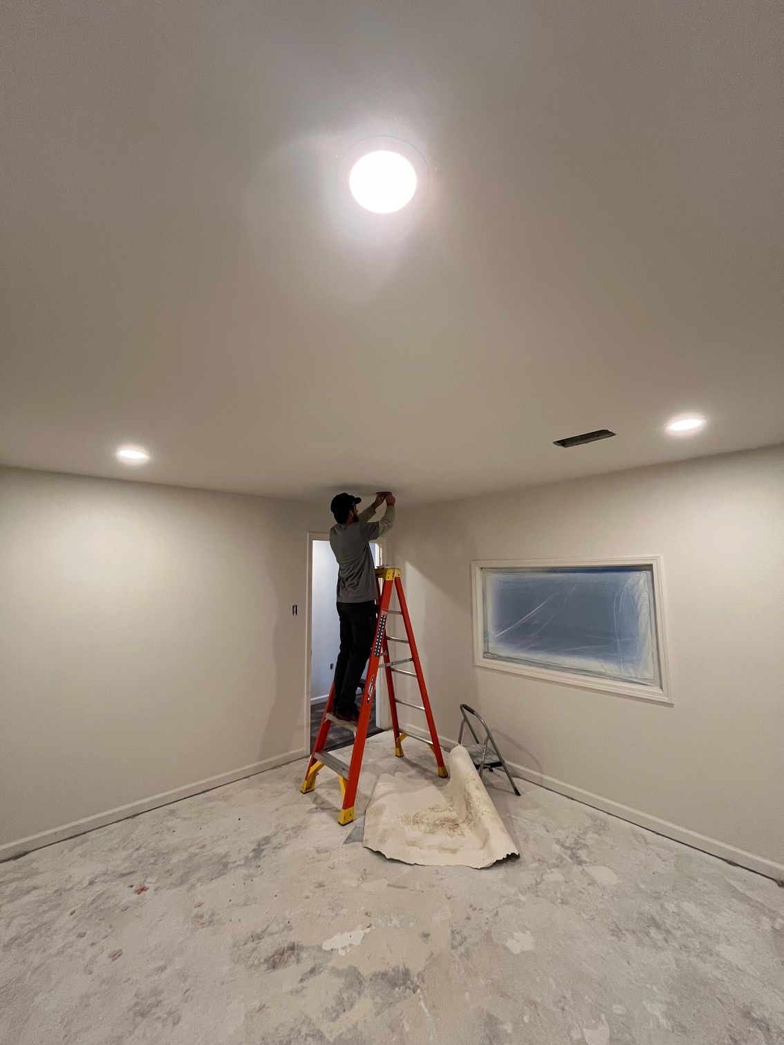 Person standing on a ladder painting a ceiling in a newly renovated room with white walls and recessed lights