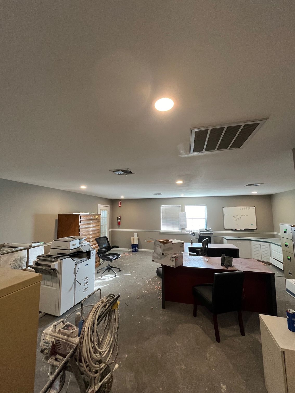 Office workspace with desks, filing cabinets, chairs, and ceiling lights in a cluttered room