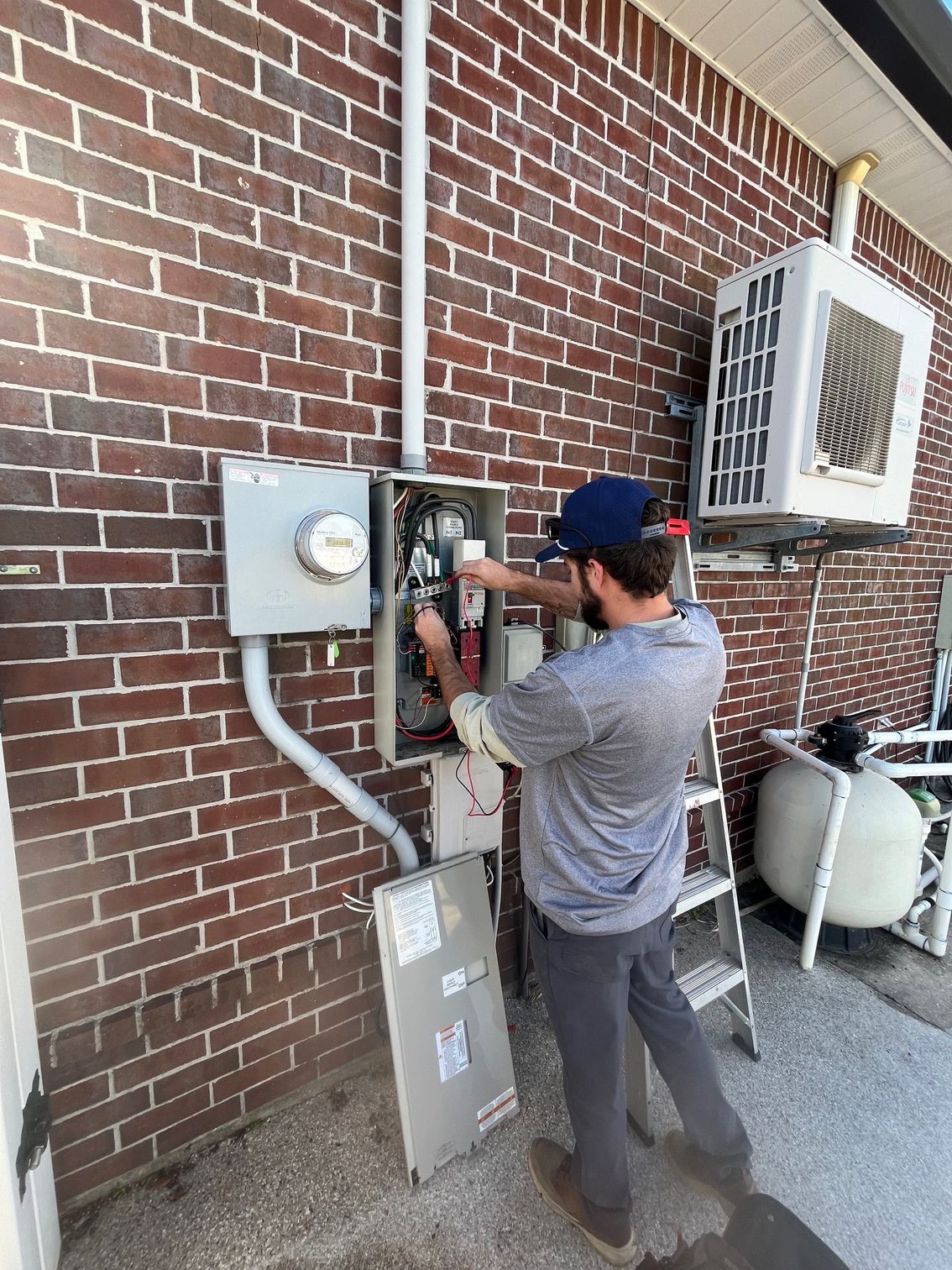 Technician inspecting electrical panels beside a brick building, with an air conditioning unit nearby