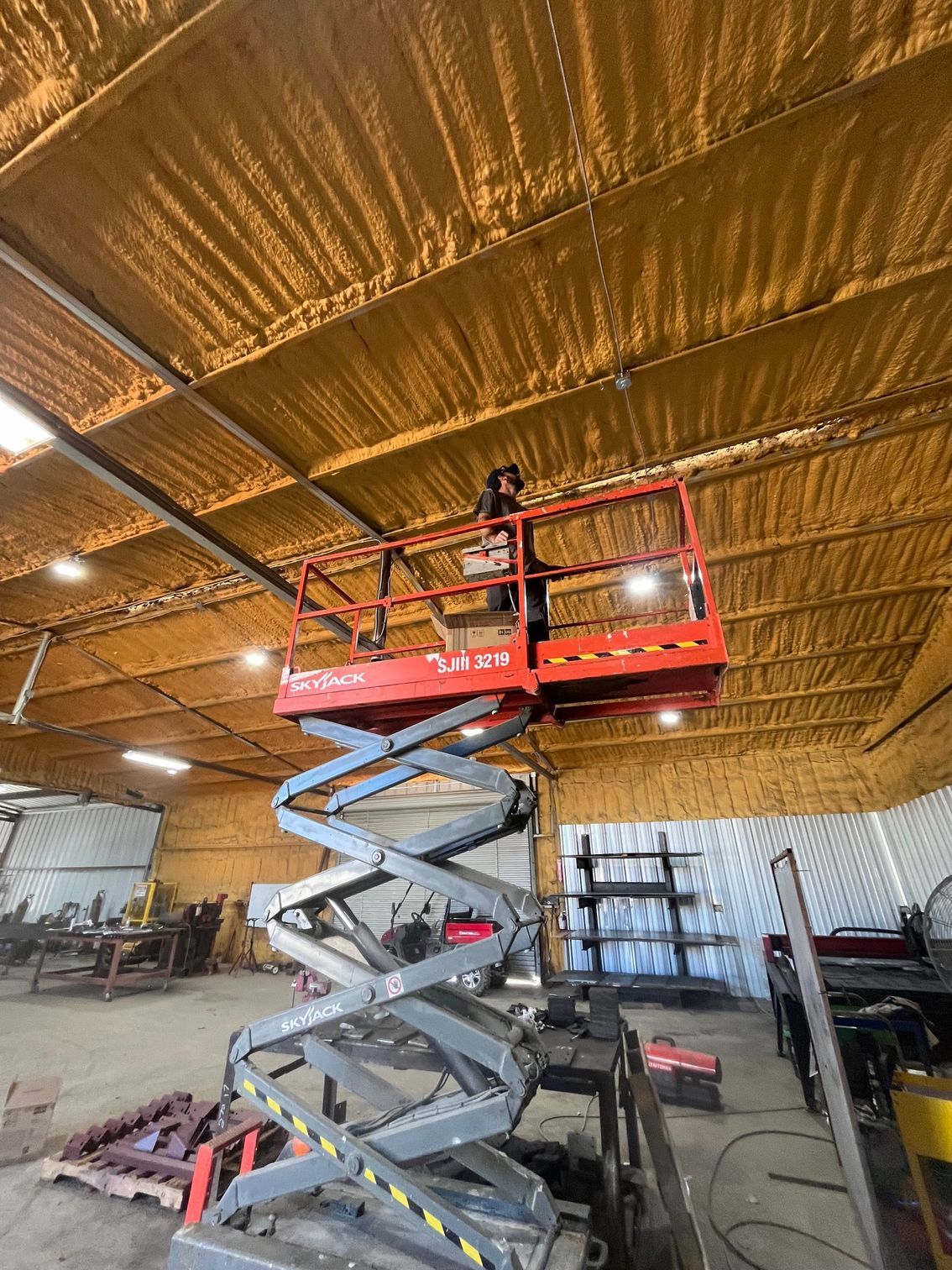 Worker in red lift basket installing ceiling insulation in a warehouse under a gold reflective roof.