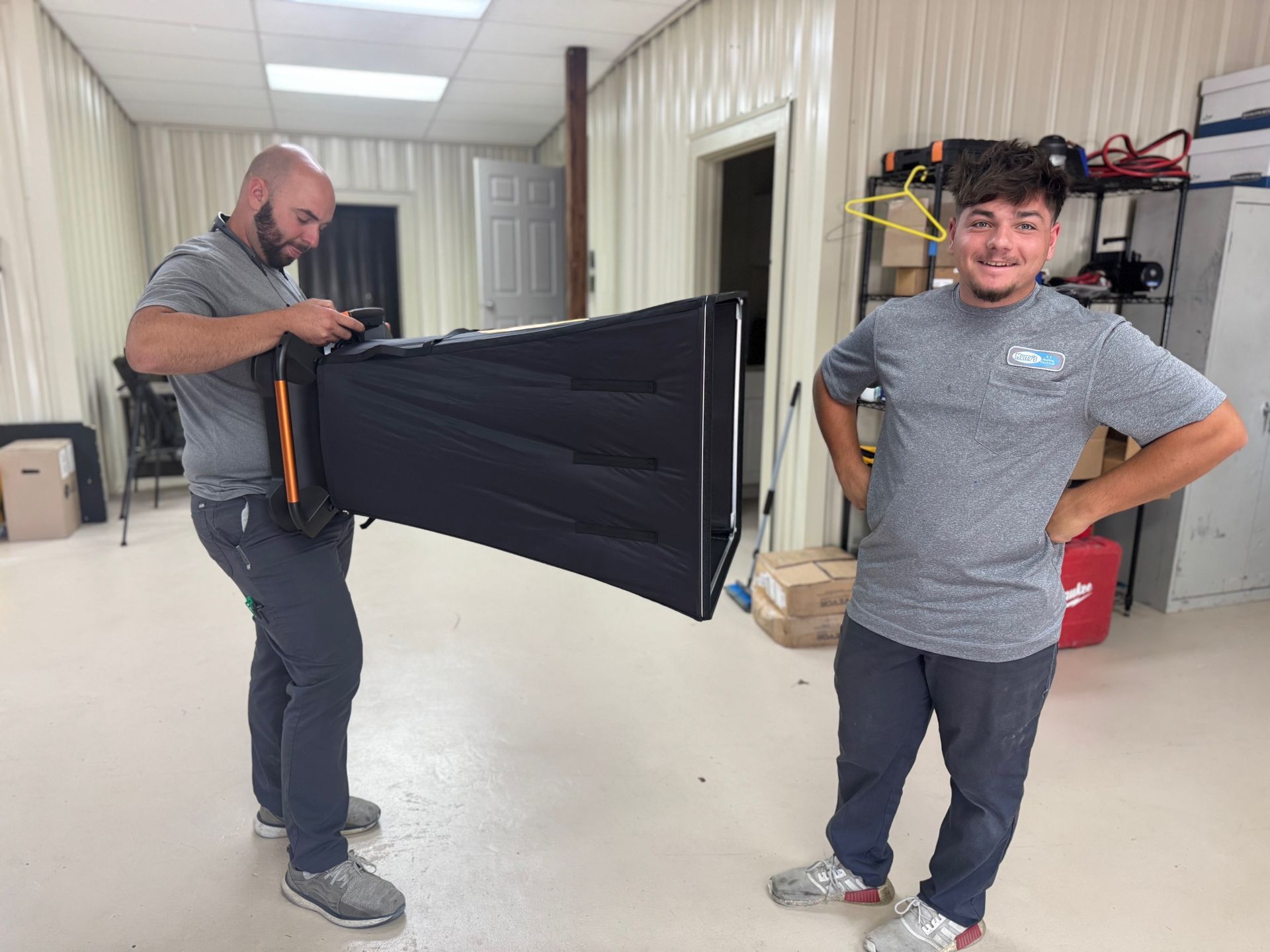 Two people in a storage room holding a large black folded item, one handling a strap while the other smiles.