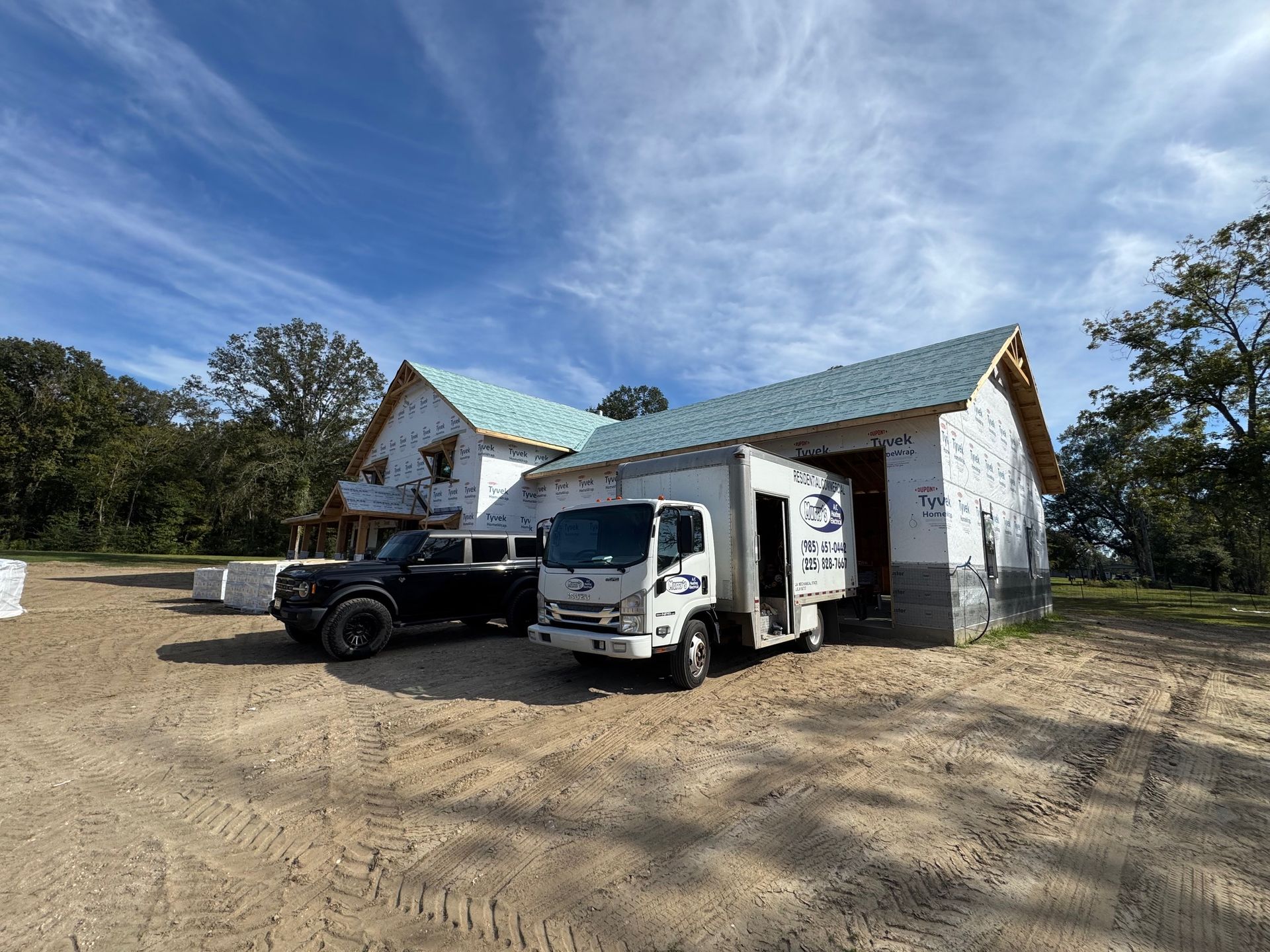White box truck and black pickup parked beside a white building with green roof on a dirt lot under a blue sky