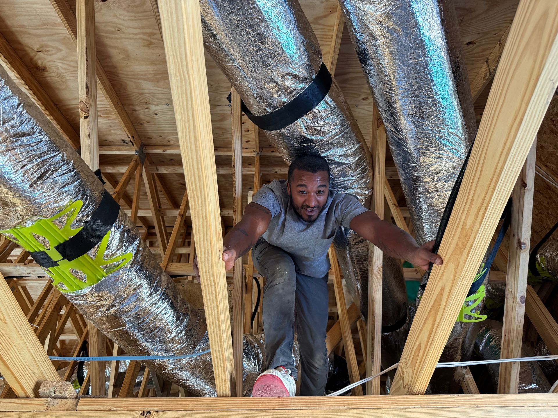 Worker crouching between wooden roof joists in an attic with silver insulated ducts overhead.