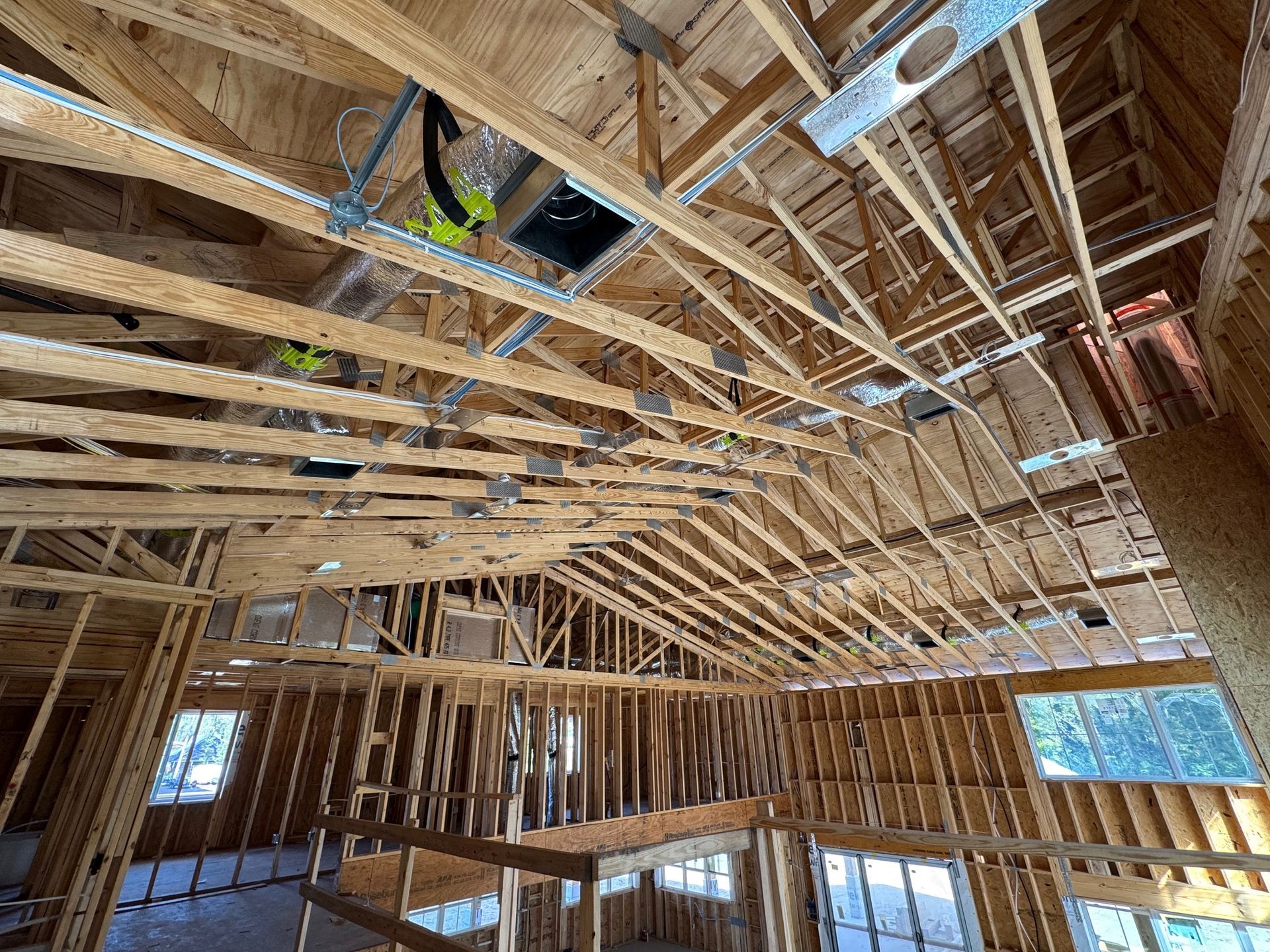 Wood-framed house interior under construction with exposed rafters, wiring, and windows showing daylight