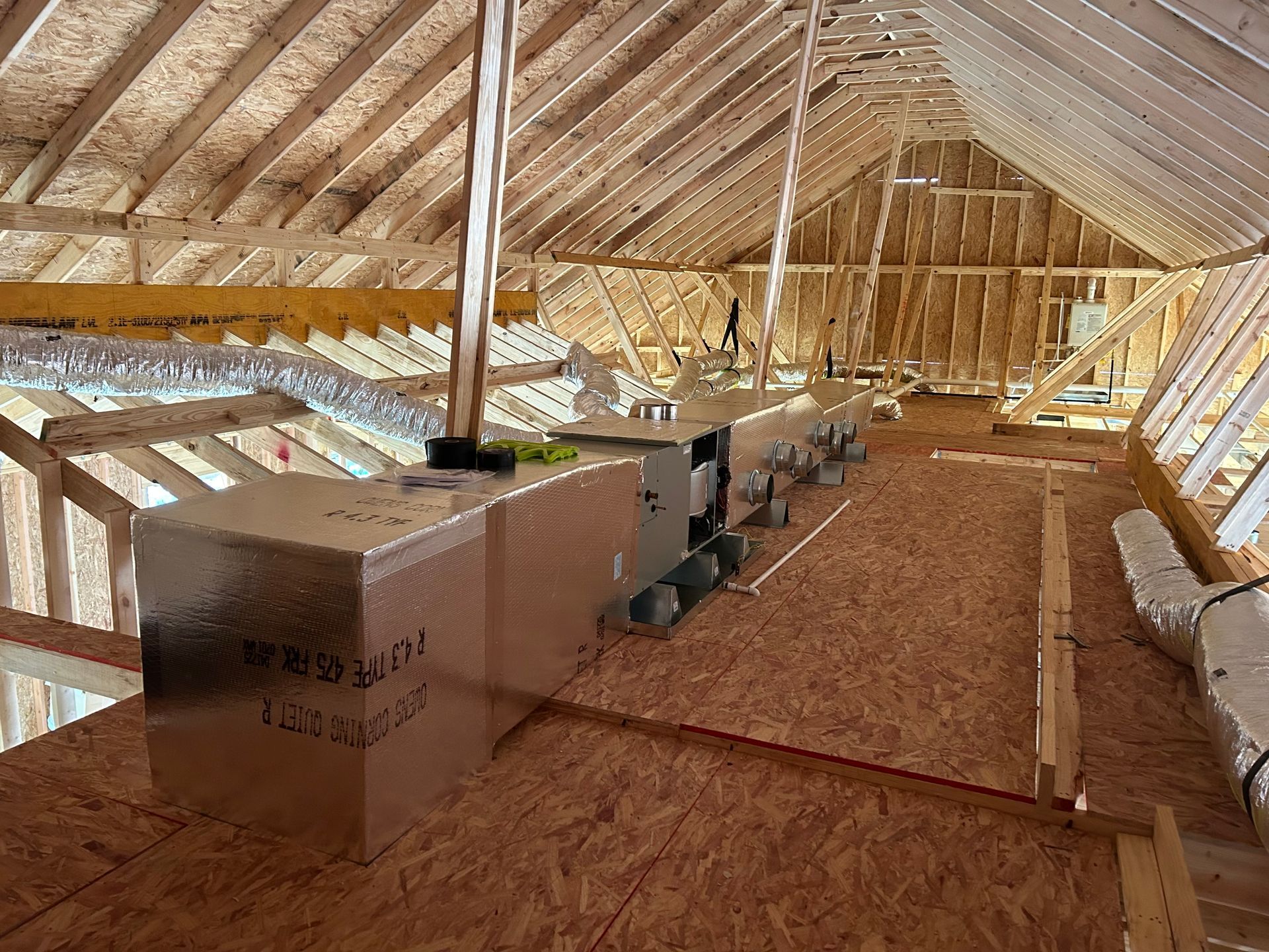 Attic with wooden rafters and a row of HVAC ducts and equipment on the floor.