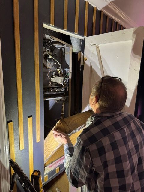 Person inspecting wiring inside an open wall panel in a room with black-and-gold striped walls.
