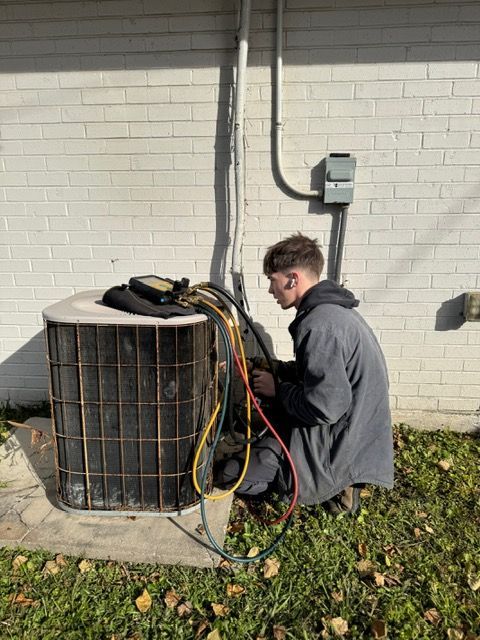 Technician kneeling beside an outdoor HVAC unit, inspecting wires against a brick wall.