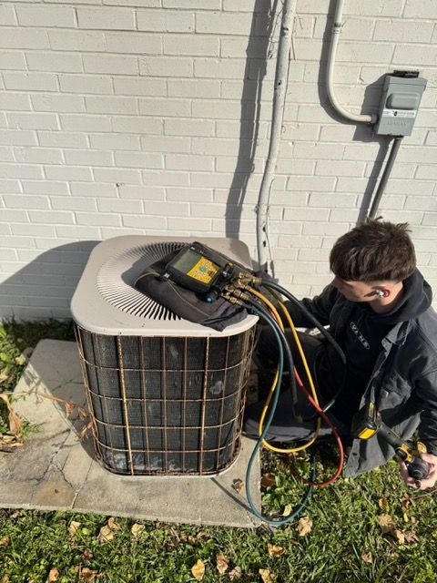 Technician kneels beside an outdoor AC unit, testing it with gauges and cables beside a house wall.