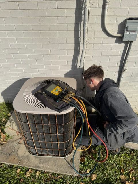 Technician servicing outdoor air conditioner unit with gauges and hoses beside a brick wall