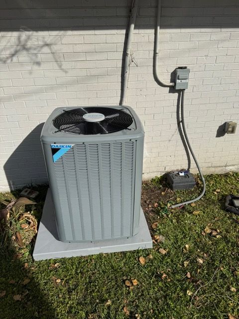 Outdoor HVAC condenser unit beside a brick wall, with a power disconnect box and grass around it.