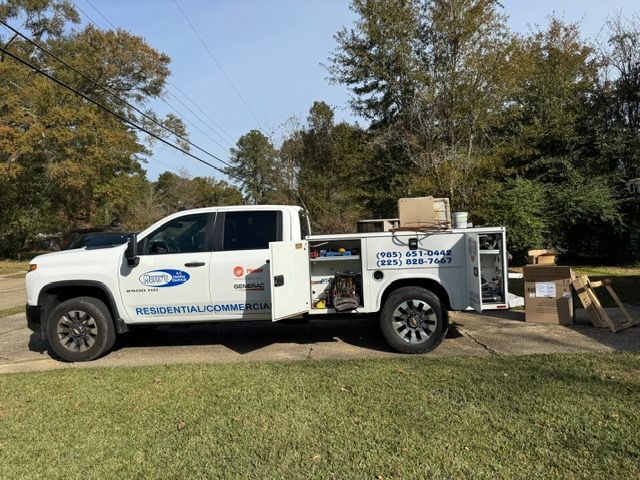 White utility truck with equipment parked on a grassy roadside beside trees and a driveway.