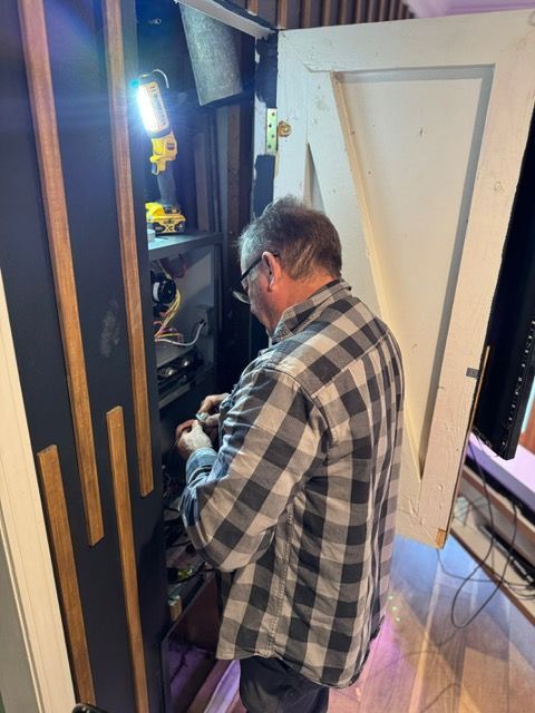 Man in plaid shirt working inside a narrow closet or utility space under bright light