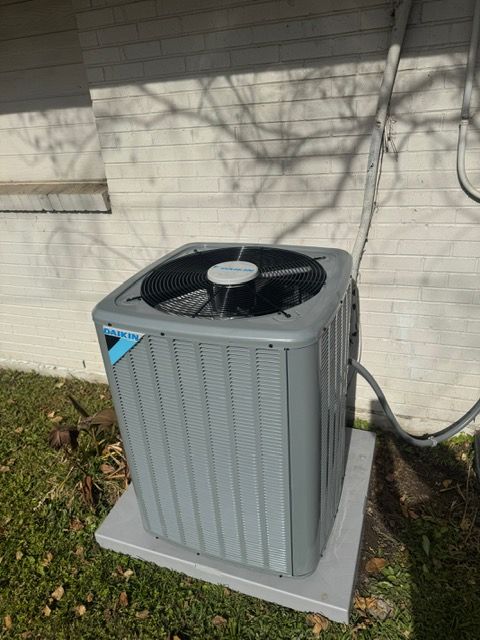 Gray outdoor air conditioner unit beside a house wall on a concrete pad, with a hose attached.