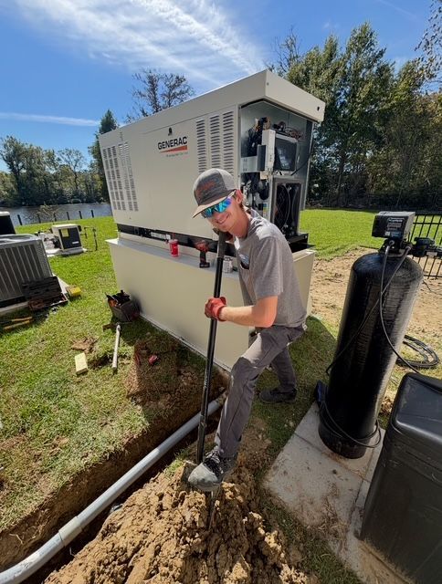 Worker shoveling dirt beside a large white utility unit outdoors on a sunny day