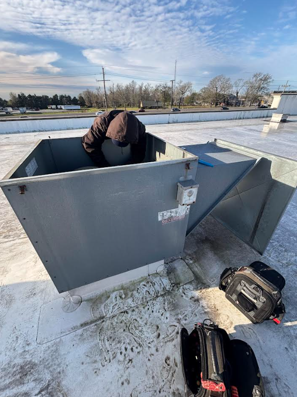 Worker bent into a large rooftop HVAC unit beside tools on a flat roof.