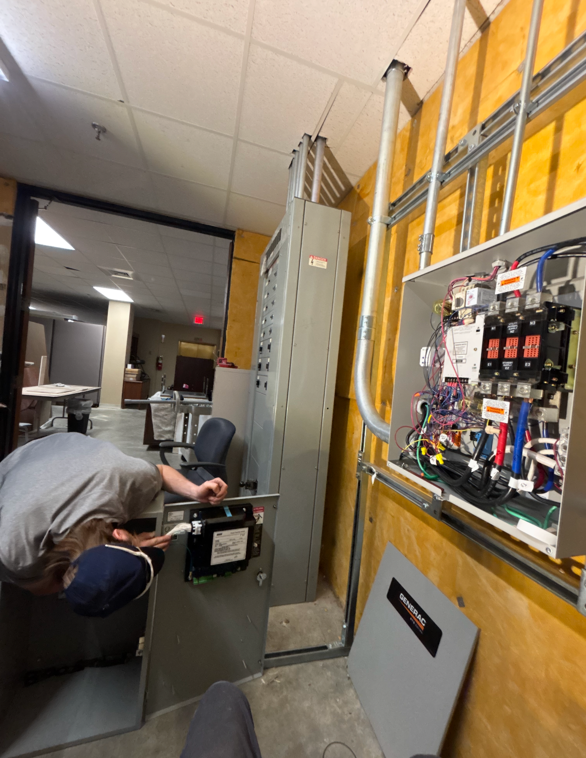 Technician working on an open electrical panel in a hallway with exposed wiring and conduit