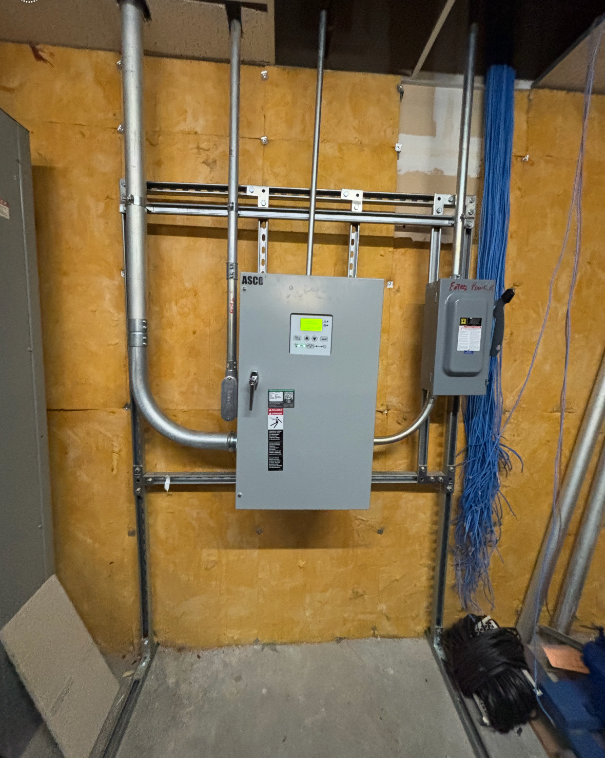 Basement utility wall with gray electrical panel, conduit, and exposed insulation.