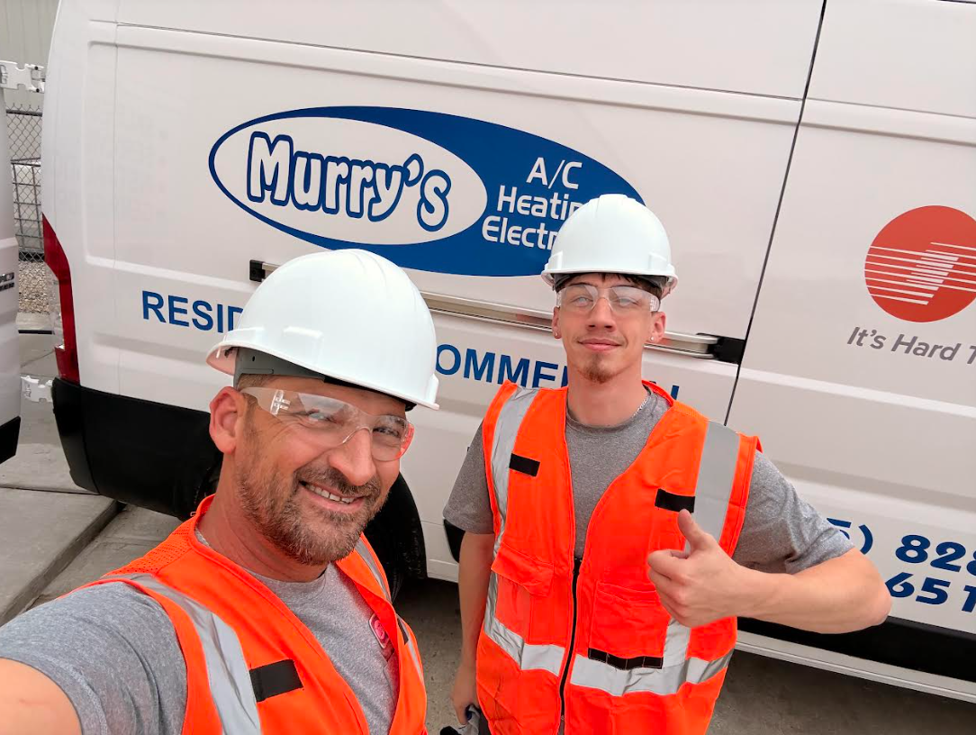 Two workers in white hard hats and orange safety vests pose beside a Murry’s HVAC service van.