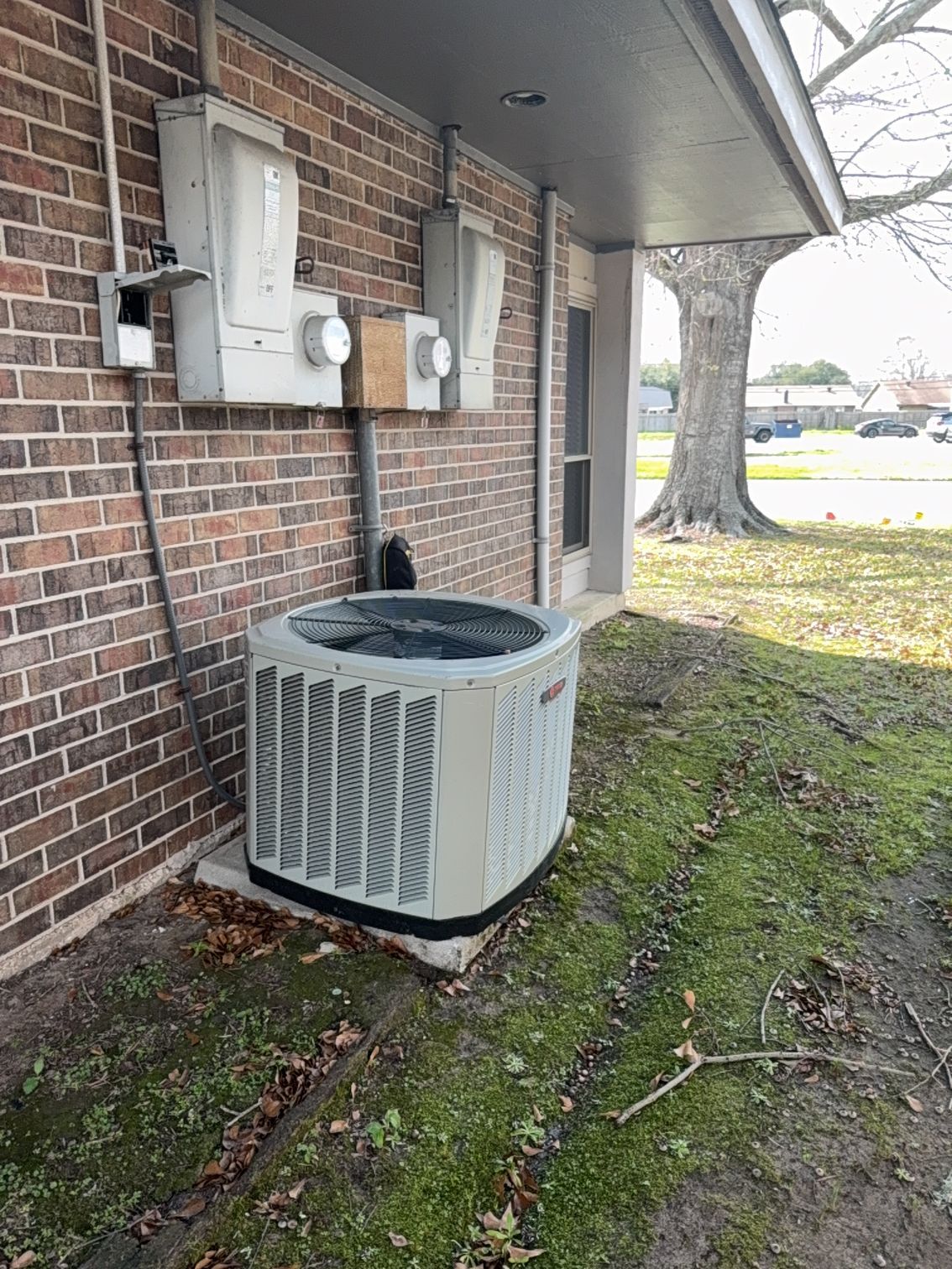 Outdoor HVAC condenser beside a brick house under an overhang, with utility boxes mounted on the wall.