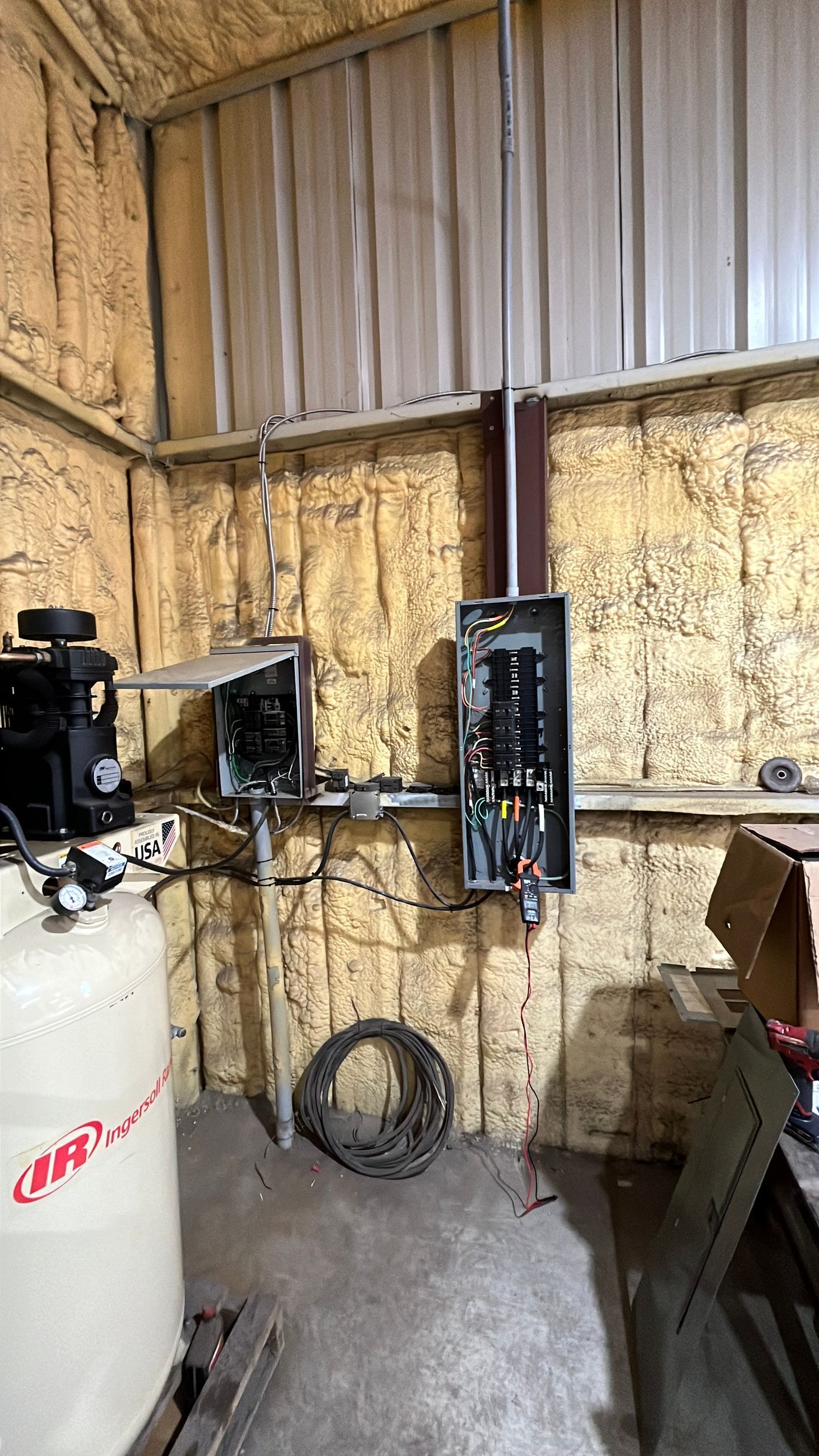 Small workshop with exposed wooden walls, electrical panel, white machine, and coiled hose on the floor.