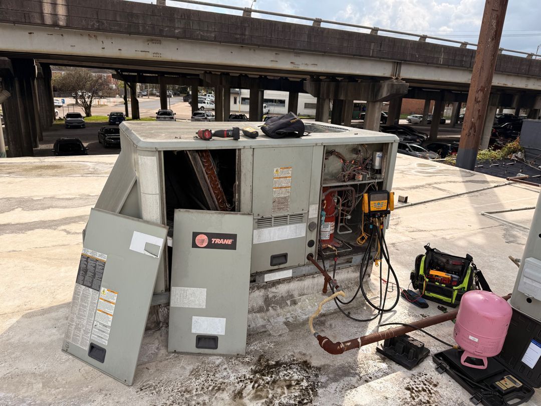 Damaged HVAC unit on a concrete pad with tools and wiring exposed under an overpass area