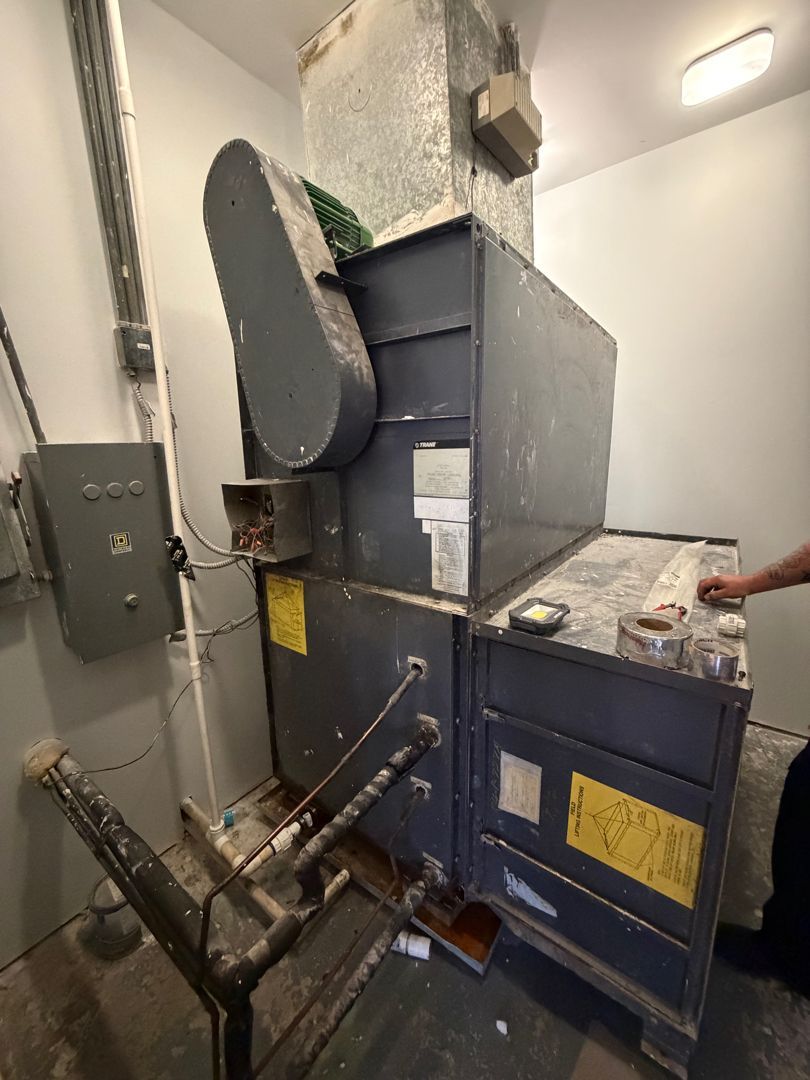 Large industrial machine in a small utility room, with gray and blue metal panels and ducts.
