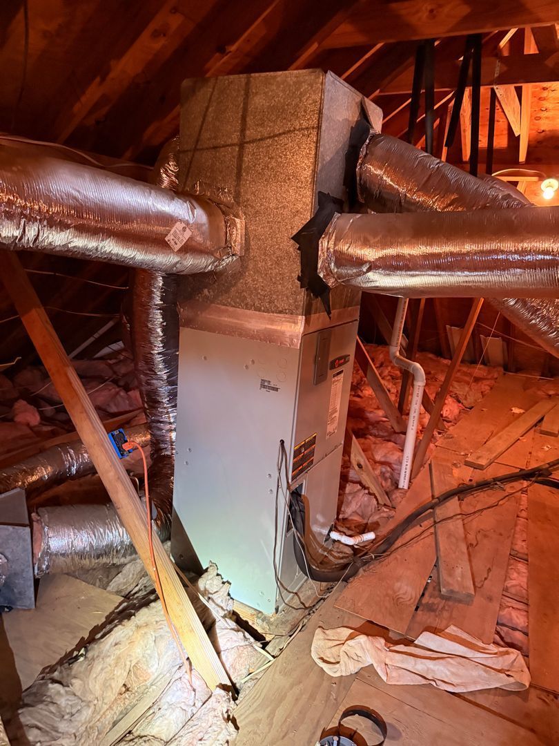 Attic HVAC ductwork around a central insulated vertical column with wooden rafters and debris on the floor