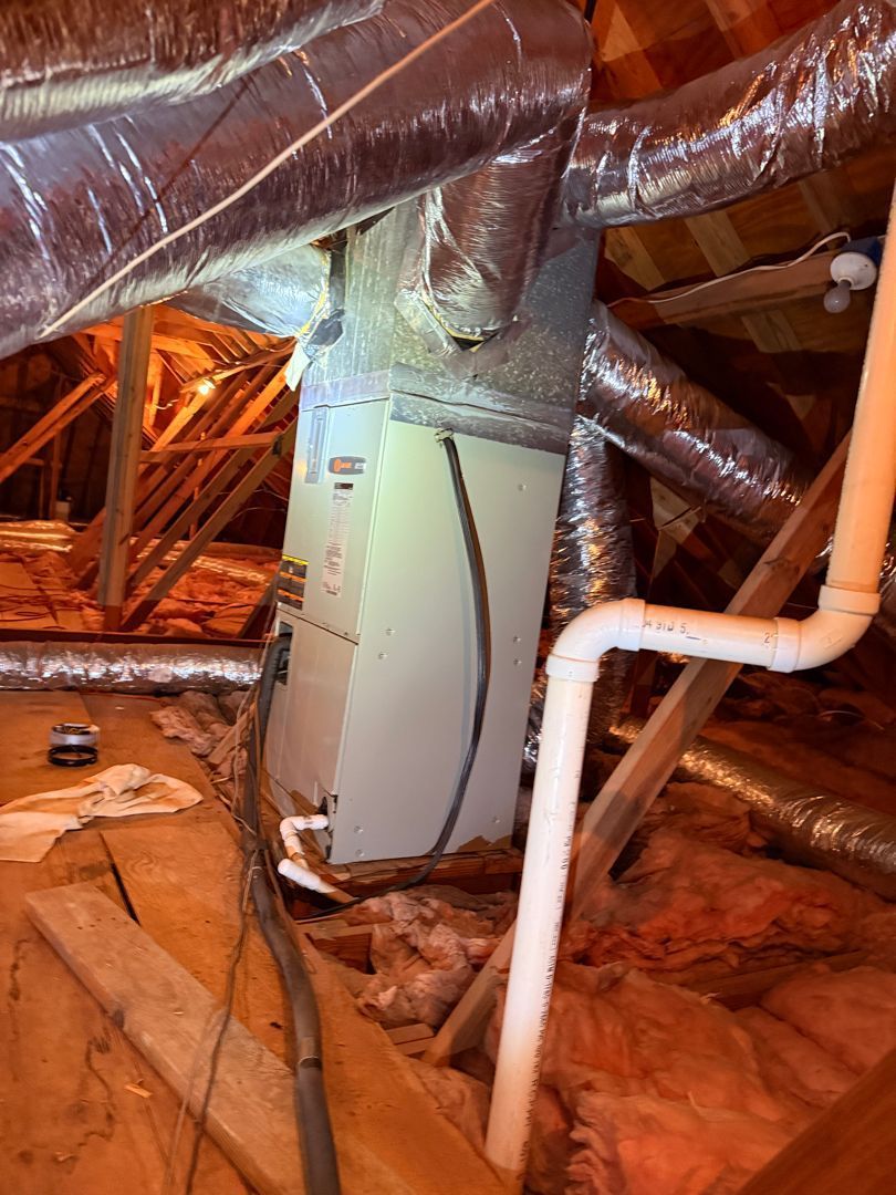 Attic HVAC unit amid ducts and white PVC pipes, surrounded by insulation and wooden rafters.