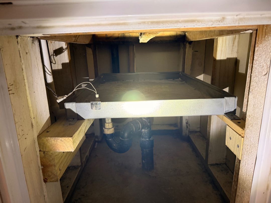 Underside of a sink with exposed pipes and a rectangular board spanning the opening, light glare in center