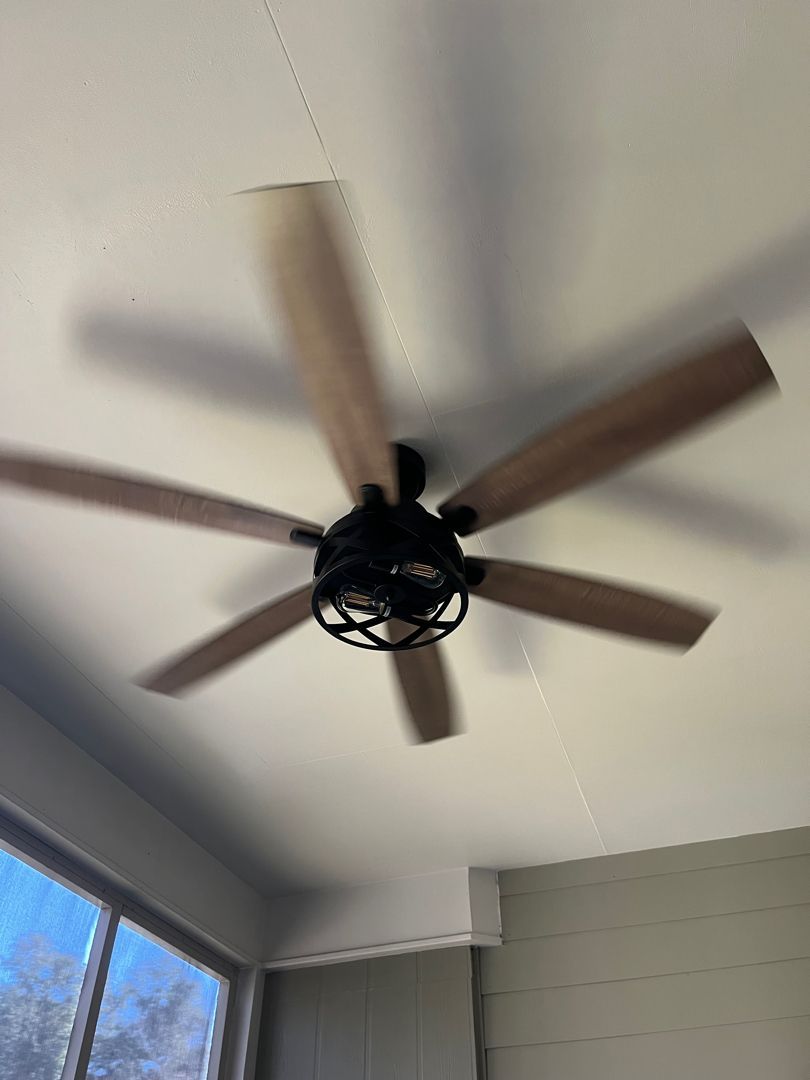 Ceiling fan with wooden blades on a white porch ceiling near a window and gray siding