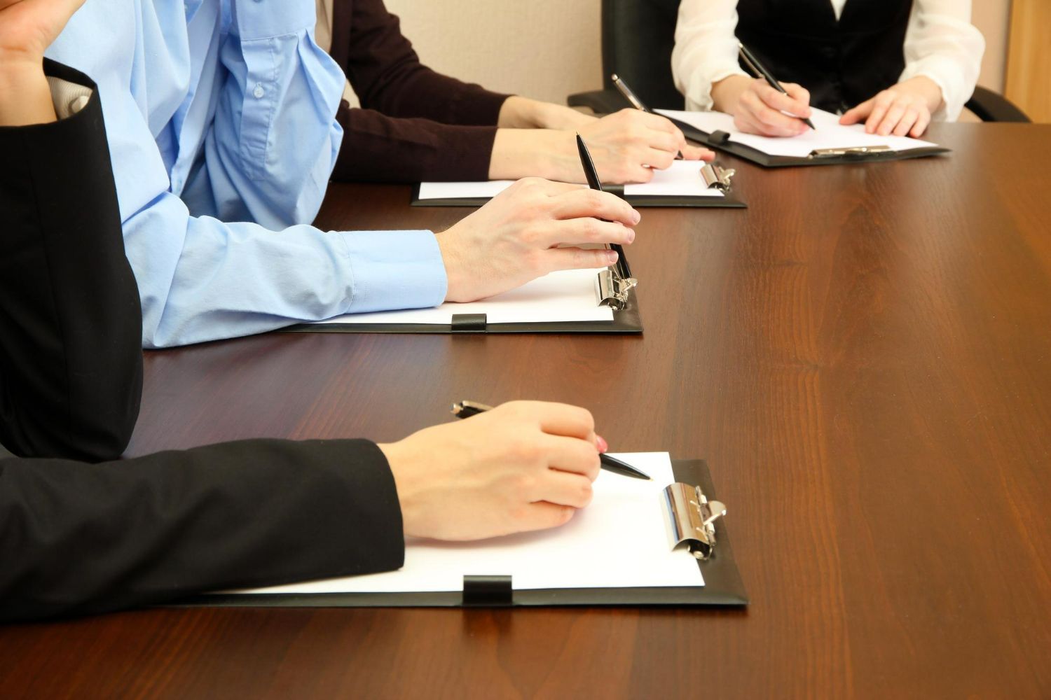 Three people sitting at a wooden table in a meeting, writing on clipboards.
