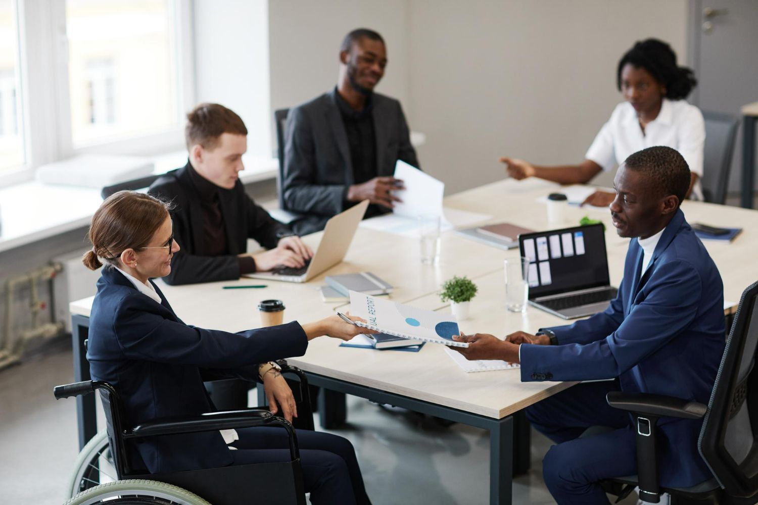 A group of professionals in a modern office meeting around a table, with one individual in a wheelchair sharing documents.