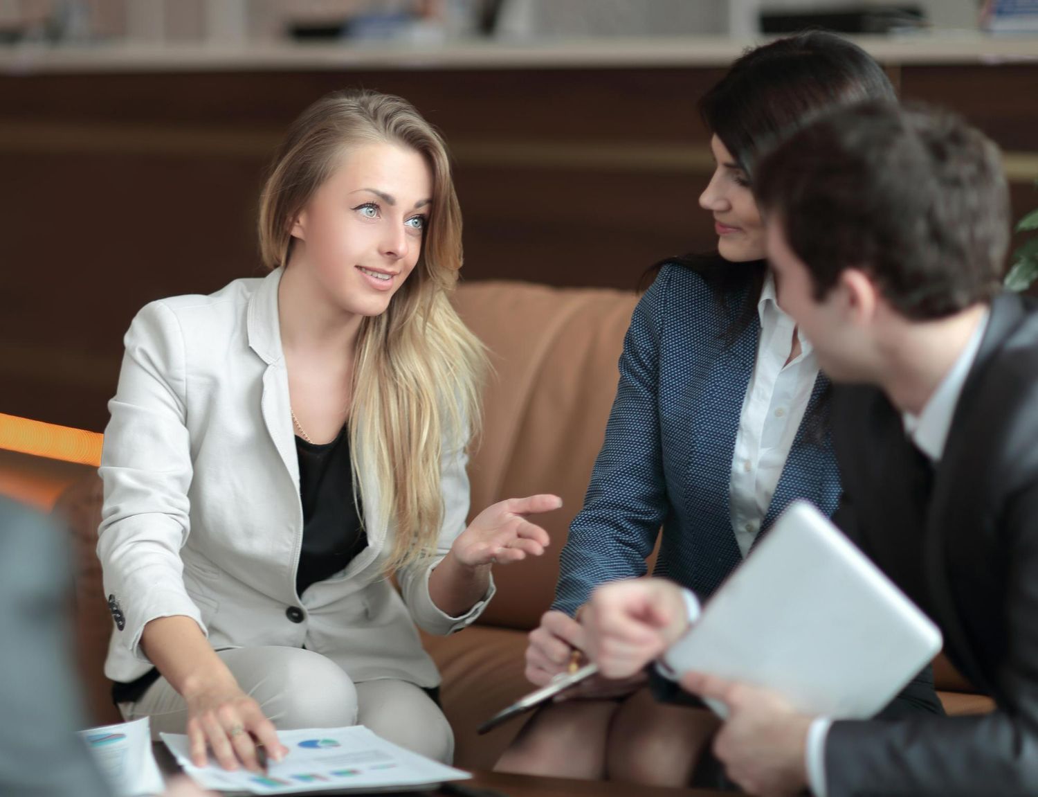 Three people in business attire sit on a sofa, discussing documents and a tablet in an office setting.