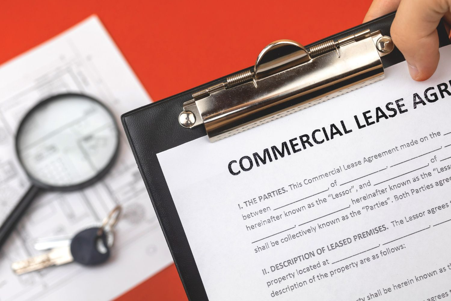 A hand holds a clipboard with a Commercial Lease Agreement over a document with a magnifying glass and keys.