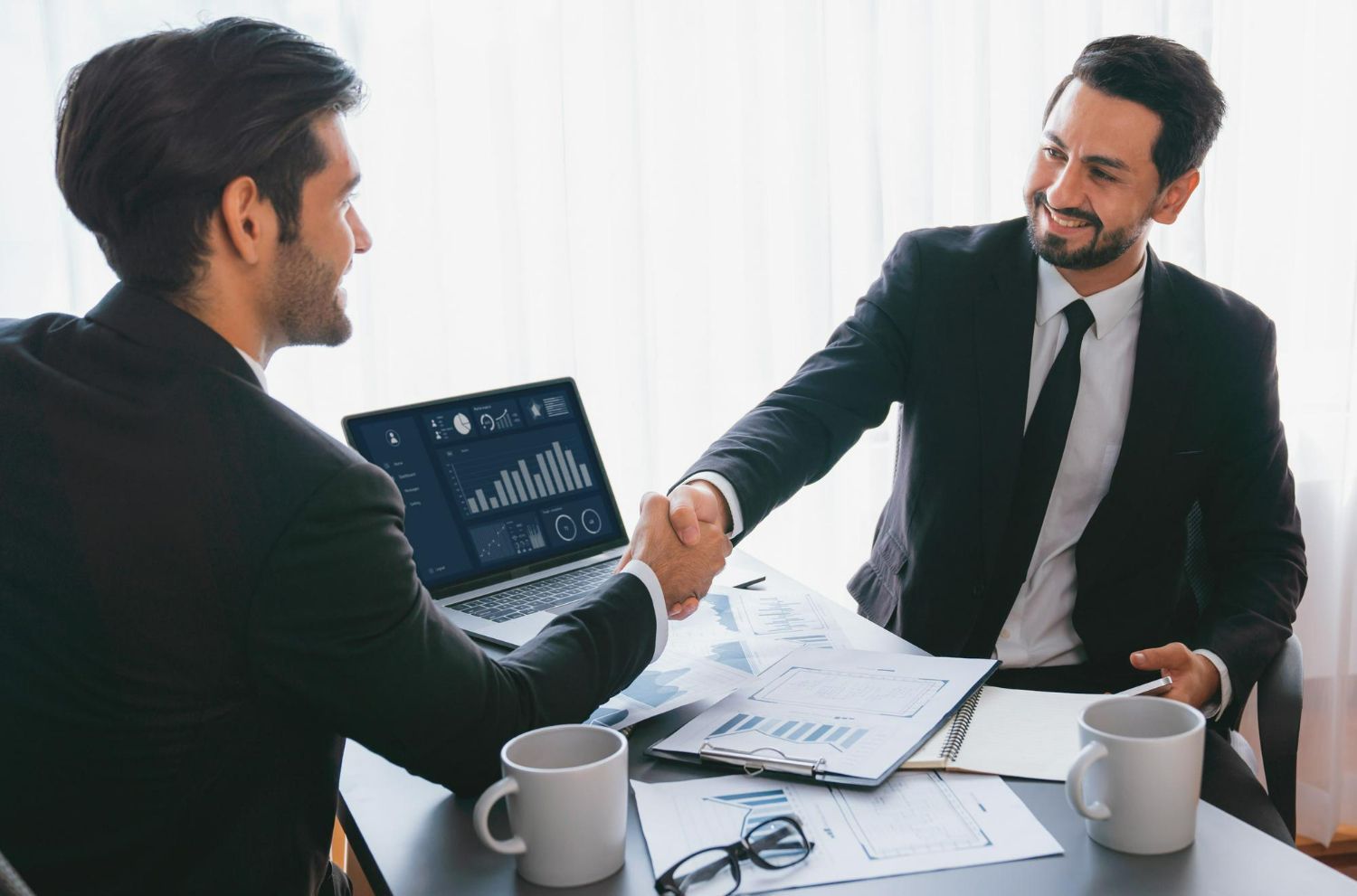 Two professionals in suits shaking hands across a desk with a laptop, documents, and coffee mugs in a bright office.