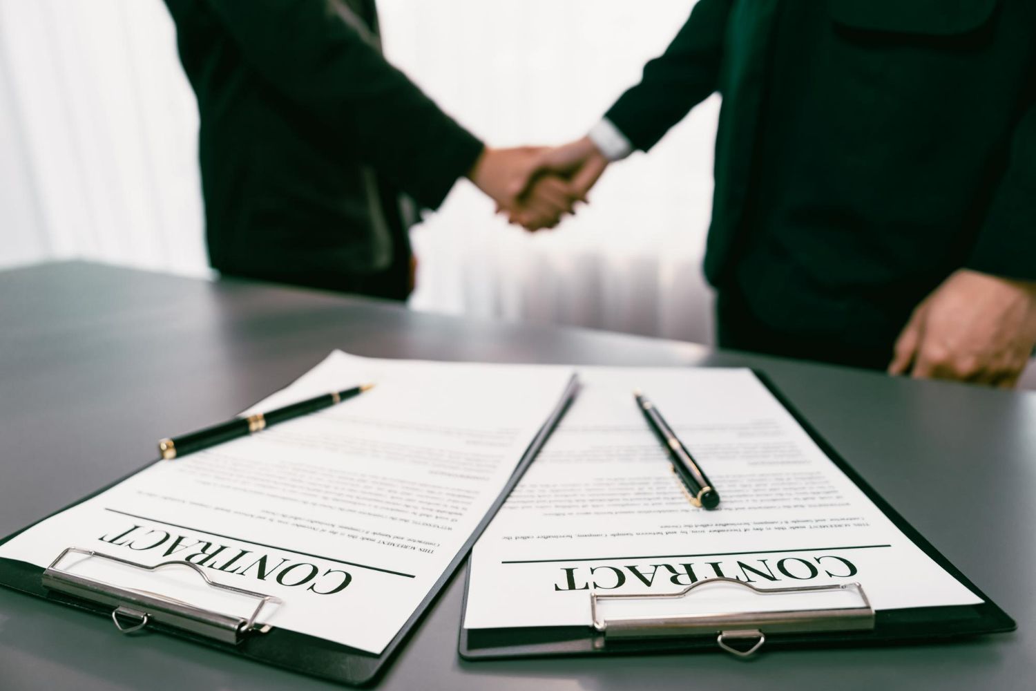 Two business people shake hands over a desk featuring two contract documents and pens.