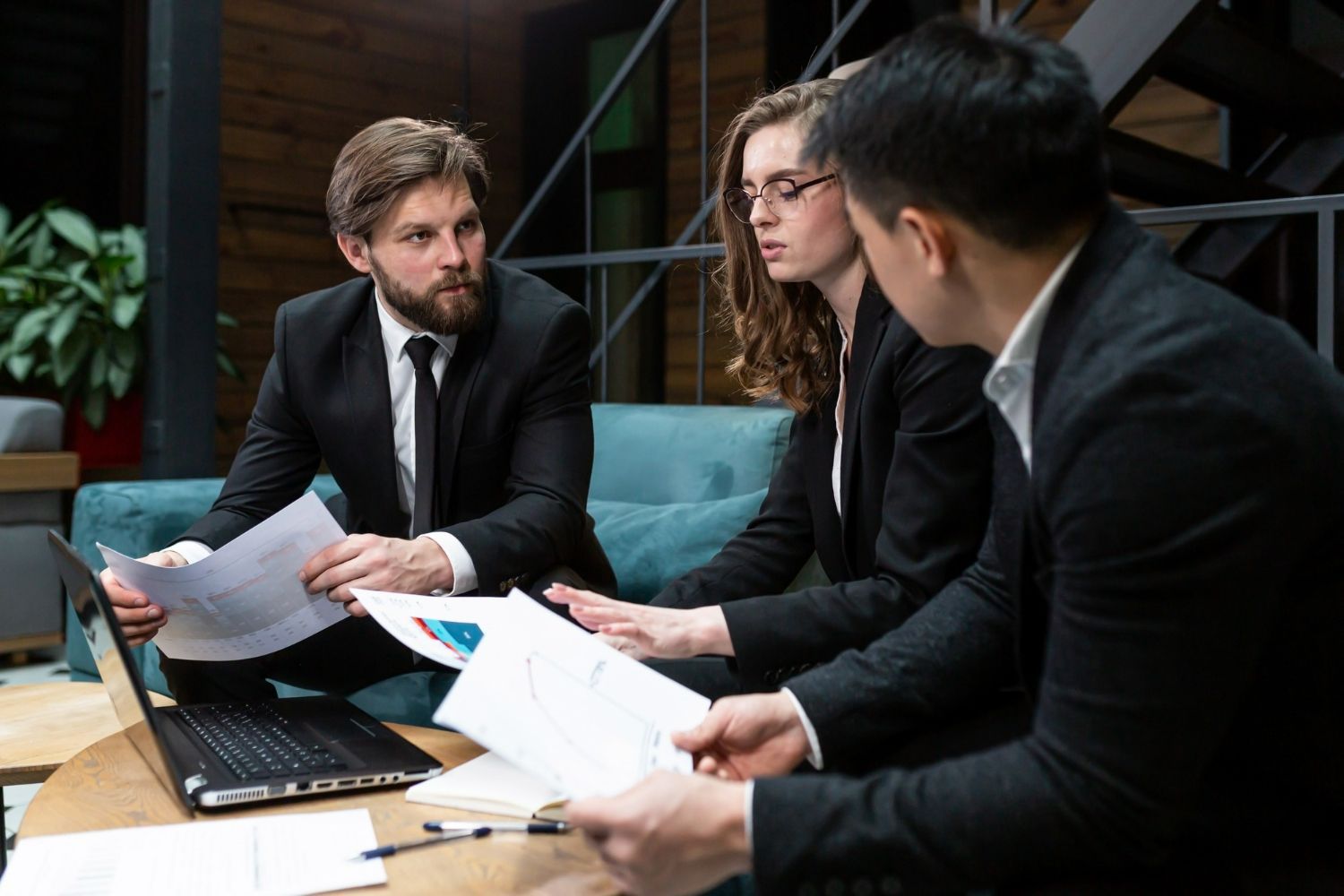 Three business professionals sitting in a modern office space, discussing documents and charts at a wooden table.