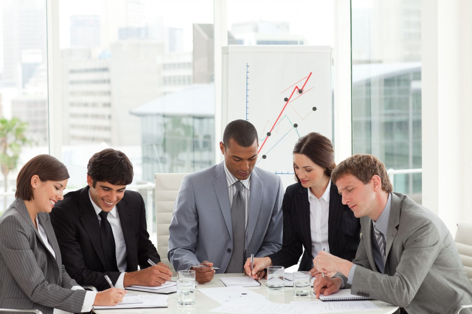 Five business professionals sit around a table in a bright office, reviewing documents with a growth chart in the background.