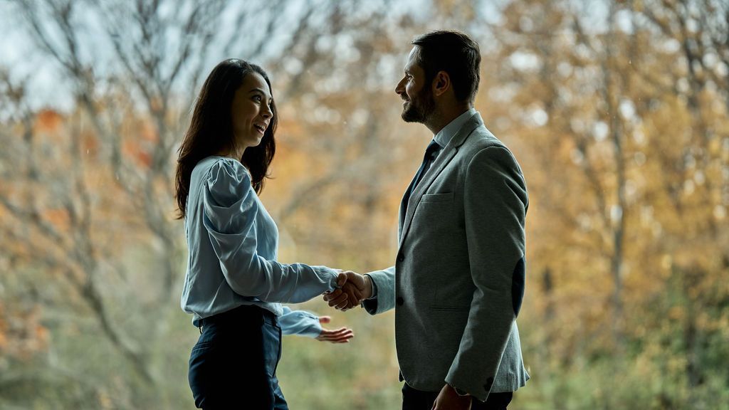 A man and a woman are shaking hands in front of a window.