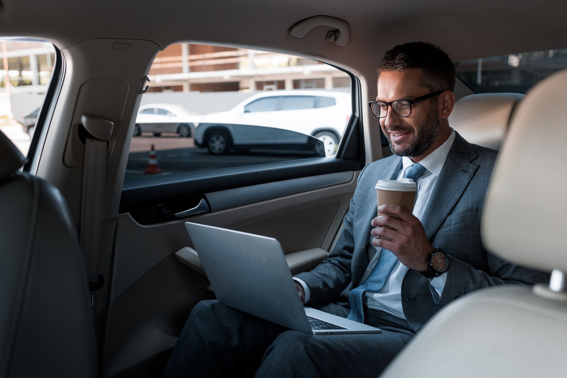 A man in a suit is sitting in the back seat of a car using a laptop and drinking coffee.